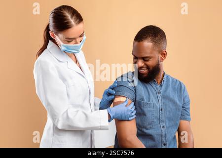 Jeune docteur en masque facial mettant un pansement sur le bras d'un patient afro-américain Banque D'Images