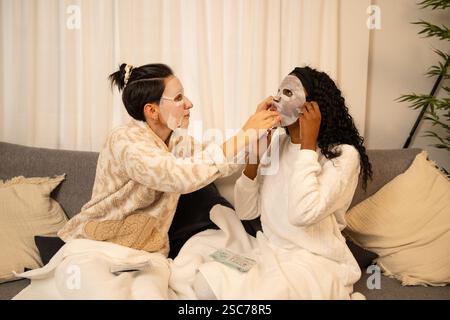 Deux amis portant des peignoirs sont assis sur un canapé dans leur salon, appliquant des masques cosmétiques en feuille sur leurs visages pour un traitement relaxant de spa à la maison Banque D'Images