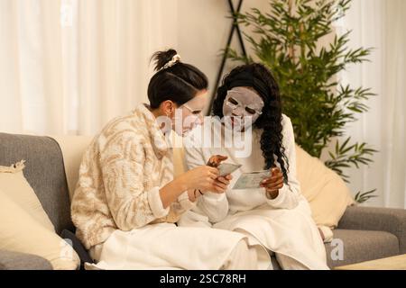 Deux femmes diverses sont assises sur un canapé portant des masques faciaux et lisant les instructions sur la façon de les appliquer, profitant d'un soin spa à domicile relaxant Banque D'Images