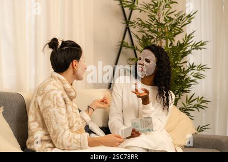Deux amies féminines diverses discutent des produits de beauté tout en portant des masques de feuille faciale et assis confortablement sur un canapé dans leur salon, enj Banque D'Images
