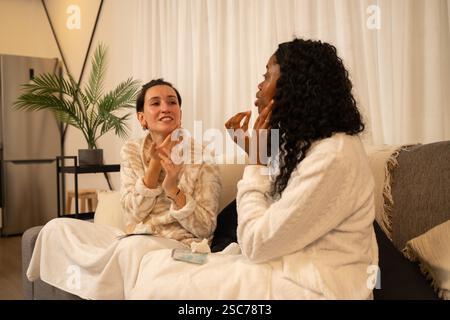 Deux femmes multiethniques portant des peignoirs sont assises sur un canapé confortable dans leur appartement, appliquant une crème pour le visage et parlant de routines de beauté Banque D'Images
