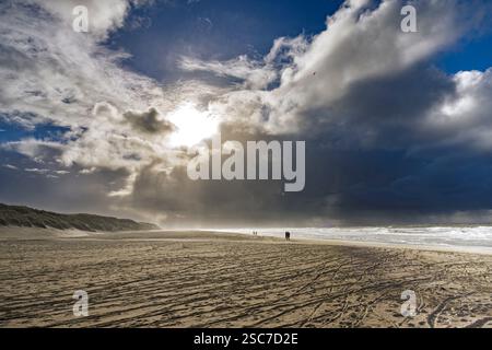 Une scène de plage dramatique avec un ciel nuageux et la lumière du soleil se perce, projetant des ombres sur le sable. Les vagues de l'océan sont visibles, et quelques personnes le sont Banque D'Images