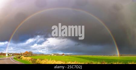 Un arc-en-ciel vibrant vole au-dessus d'un paysage verdoyant sous un ciel spectaculaire rempli de nuages sombres. La scène capture le contraste entre le rai coloré Banque D'Images