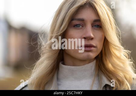 Une jeune femme se tient dehors avec une expression sereine, mettant en valeur ses longs cheveux blonds et portant un pull de couleur claire. La lumière douce du soleil s'allume Banque D'Images
