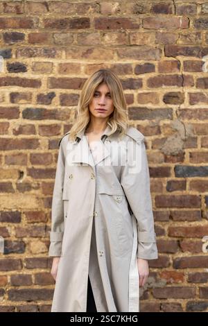 Une jeune femme se tient confiante dans un trench-coat beige, mettant en valeur son sens de la mode contre un mur de briques rustique. La lumière naturelle met en valeur la scène, Banque D'Images