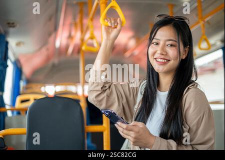 Une belle et attrayante femme asiatique dans des vêtements décontractés est dans un bus, tenant une main courante et souriant à la caméra. transports publics, style de vie, ci Banque D'Images