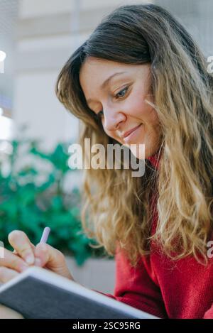 Jeune femme blonde portant un pull rouge à col roulé écrit des notes dans un cahier à l'aide d'un stylo, souriante et paraissant concentrée en position assise Banque D'Images
