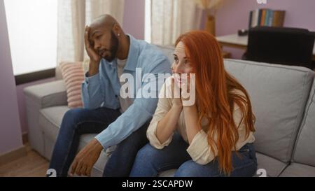 Couple interracial assis à l'intérieur sur un canapé à la maison regardant préoccupé montrant des expressions émotionnelles dans un intérieur de salon Banque D'Images