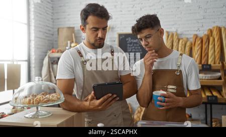 Deux boulangers masculins hispaniques dans une boulangerie portant des tabliers travaillent ensemble, l'un tenant une tasse de café tandis que l'autre utilise une tablette, entourée de f Banque D'Images