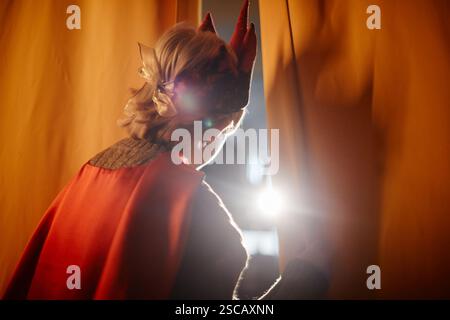 Personne portant un costume de scène regardant à travers les rideaux, illuminée par une lumière vive. Capturer un moment dramatique lors d'une représentation théâtrale Banque D'Images