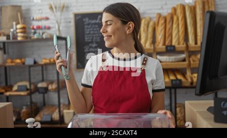 Jeune femme dans un tablier rouge travaille dans une boulangerie en utilisant des pinces de cuisine entourées d'étagères de pain frais et de pâtisseries Banque D'Images