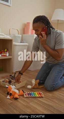 Jeune homme avec des tresses agenouillant dans le salon tout en parlant au téléphone et en rangeant les jouets améliore une atmosphère chaleureuse à la maison. Banque D'Images