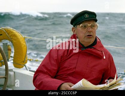 Un homme avec un anorak rouge, des lunettes et une casquette regarde une carte nautique au large de la Norvège en haute mer. [traduction automatique] Banque D'Images