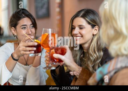 Gros plan de trois jeunes femmes élégantes qui grillent avec des cocktails colorés dans un bar branché, profitant d'une soirée animée avec des amis. Banque D'Images