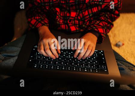 Vue en angle élevé d'une fille d'enfant méconnaissable portant une chemise à carreaux rouge et noir tapant sur un clavier d'ordinateur portable rétroéclairé assis sur le lit avec les jambes croisées Banque D'Images