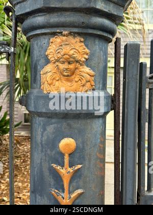Vue détaillée d'une porte en fer ornée à la Nouvelle-Orléans, avec un relief unique d'un visage entouré de plantes tropicales Banque D'Images