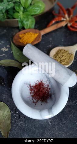 Une petite quantité de vrai safran rouge dans un mortier de marbre gris clair. En arrière-plan - épices, feuilles de laurier, piment et basilic. Banque D'Images