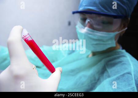 Main gantée présentant un tube à essai rempli de liquide rouge, avec un individu masqué en gommage observant Banque D'Images