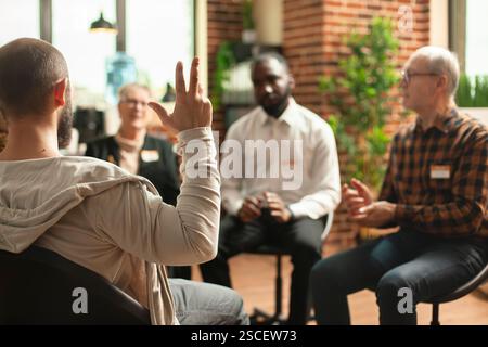 Focalisation sélective sur le jeune homme faisant des gestes avec sa main pendant la séance de thérapie. Un groupe de personnes diverses est assis dans une salle de mur de briques, discutant des luttes de santé mentale et des progrès. Banque D'Images