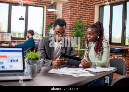Homme d'affaires expliquant la stratégie de marketing sur tablette à une collègue féminine dans l'espace de travail de mur de briques. Collègues afro-américains assis à table avec des documents et ayant une conversation sérieuse au bureau. Banque D'Images