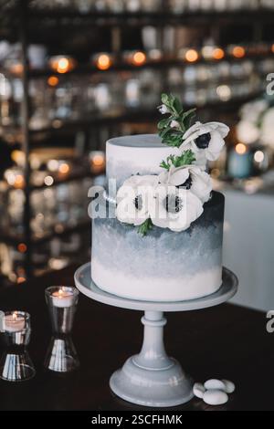 Un gâteau de mariage à deux niveaux magnifiquement conçu se tient sur un stand de gâteau gris, orné de délicates fleurs blanches. L'arrière-plan dispose d'un cadre confortable Banque D'Images