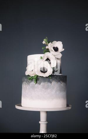 Un superbe gâteau à deux niveaux dispose d'un motif d'ombre gris lisse, orné de fleurs blanches réalistes avec des centres sombres, affichés élégamment sur un simple Banque D'Images