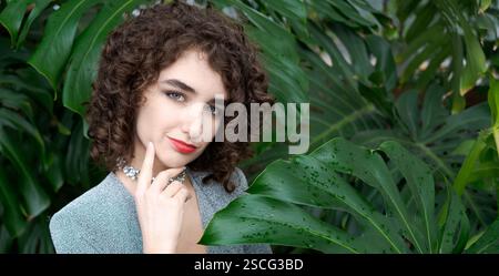 Une femme aux cheveux bouclés se tient devant une plante verte. Elle porte une robe bleue et un collier Banque D'Images