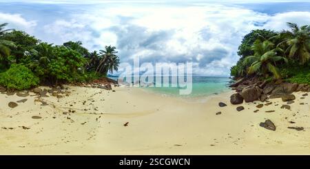 Une plage tranquille avec des rochers éparpillés et des eaux turquoises, entourée d'arbres tropicaux luxuriants sous un ciel nuageux. Plage de l'anse Bougainville, Seychelles. panorama 360. Banque D'Images