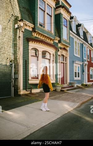 Vue arrière d'une jeune femme méconnaissable alors qu'elle marche dans une rue pittoresque bordée de maisons aux couleurs vives à l'endroit de John's, Terre-Neuve-et-Labrador, Banque D'Images