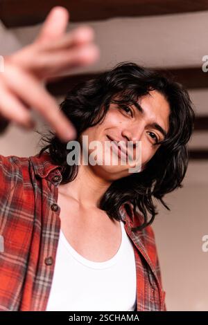 Un jeune homme souriant dans un débardeur blanc et une chemise à carreaux tend sa main vers l'appareil photo, donnant un geste amical et invitant dans un chaleureux éclairage Banque D'Images