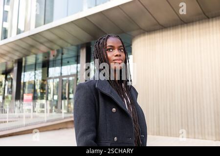 Une jeune femme d'affaires afro-américaine professionnelle avec de longues tresses à l'extérieur, regardant loin confiant dans un cadre d'affaires. Banque D'Images