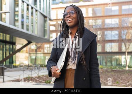 Une femme d'affaires afro-américaine aux cheveux tressés souriant et tenant un ordinateur portable tout en prenant une pause à l'extérieur près d'un immeuble de bureaux moderne. Banque D'Images