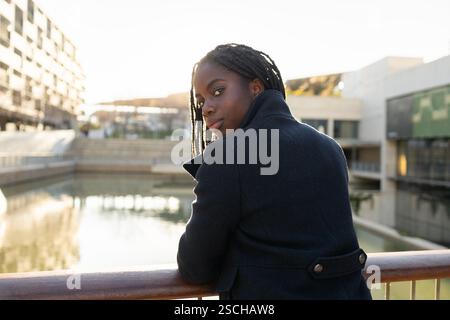 Femme d'affaires afro-américaine avec des tresses prend une pause rafraîchissante à l'extérieur, s'appuyant sur une balustrade avec un fond d'eau sereine et des bâtiments urbains. Banque D'Images