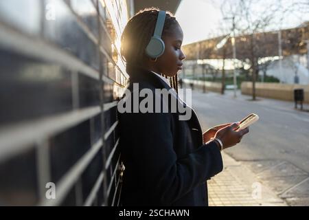 Une femme d’affaires afro-américaine, au sport des tresses et des écouteurs, est concentrée sur son smartphone lors d’une pause à l’extérieur, illuminée par un doux coucher de soleil Li Banque D'Images