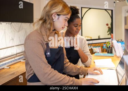 Deux employés de magasin de cosmétiques travaillant ensemble, concentrés sur un écran d'ordinateur portable. Ils portent des tabliers, entourés de fournitures de bureau, démontrant l'équipe Banque D'Images
