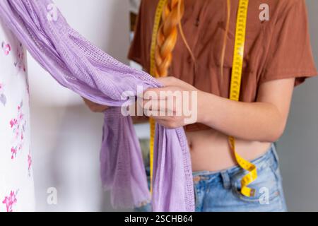 Une jeune femme examine une écharpe violette, organisant son placard pour vendre des vêtements elle porte une tenue décontractée et du ruban à mesurer autour de son cou, soigneusement Banque D'Images