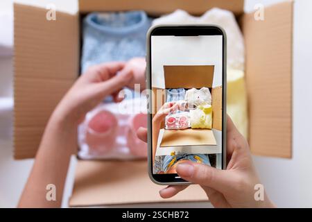 Une jeune femme utilise son smartphone pour photographier des vêtements pour une vente en ligne. Elle tient le téléphone tout en organisant les vêtements dans une boîte, capturant EA Banque D'Images
