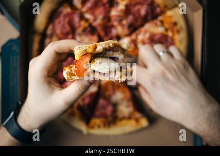 Vue d'en haut, vue rapprochée méconnaissable de mains partageant des tranches d'une pizza au fromage chaude garnie de pepperoni et de saucisse. Un rassemblement confortable avec fri Banque D'Images