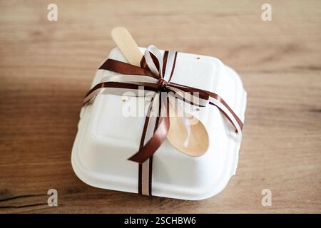 Gâteau d'anniversaire bento élégamment emballé dans une boîte à emporter blanche ornée d'un ruban chocolat-brun et d'une cuillère en bois, idéal pour les cadeaux. Banque D'Images