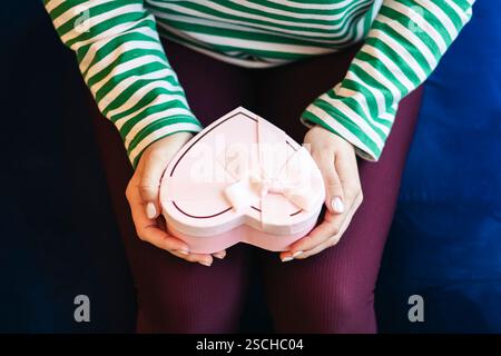 Vue de dessus recadrée femme méconnaissable dans une chemise rayée tenant une boîte cadeau rose en forme de coeur. Concentrez-vous sur les mains et la boîte, gros plan détaillé. Banque D'Images