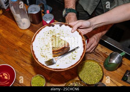 Une tranche plaquée de cheesecake surmontée de pistaches écrasées est présentée sur une assiette rustique avec une fourchette. L'arrière-plan montre un kitche de café occupé Banque D'Images
