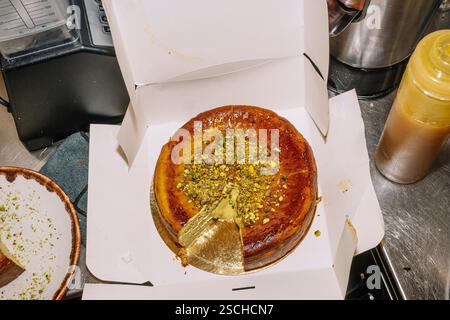 Un gâteau au fromage luxueux garni de pistaches écrasées et de feuilles d'or se trouve élégamment dans une boîte ouverte. La scène se déroule dans un environnement de boulangerie moderne. Banque D'Images
