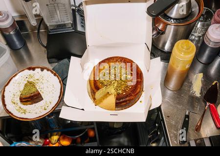 Un cheesecake fraîchement cuit garni de pistaches écrasées se trouve dans une boîte ouverte, avec une tranche plaquée à proximité. La scène capture une cuisine de café wi Banque D'Images