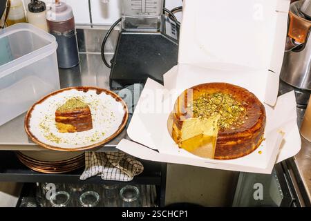 Un riche cheesecake garni de pistaches écrasées se trouve dans une boîte ouverte, avec une tranche servie dans une assiette à proximité. La scène se déroule dans une cuisine de café Banque D'Images