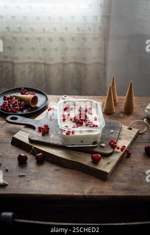 Scène rustique de crème glacée à la vanille maison garnie de groseilles rouges et de baies, servie sur une planche de bois avec des cerises éparpillées. Banque D'Images
