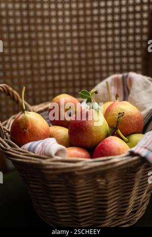 Un charmant panier en osier rempli de poires mûres, reposant sur un textile rayé. Les tons chauds et la texture douce créent une ambiance confortable et rustique parfaite Banque D'Images