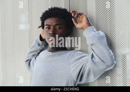 Un jeune homme noir élégamment habillé d'un sweat-shirt gris pose avec confiance contre un mur blanc texturé numériquement, regardant l'appareil photo. Banque D'Images