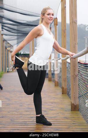 Une jeune femme blonde, vêtue de vêtements de sport, s'étire sur une passerelle en bois, affichant un style de vie sain et un esprit sportif. Banque D'Images