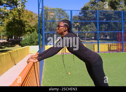 Une joueuse de basket-ball dans une tenue noire effectue un exercice d'étirement dans un parc coloré. Le ciel bleu clair et l'environnement vert améliorent la v Banque D'Images