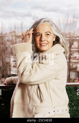 Femme âgée souriante avec les cheveux blancs se tient sur son balcon, portant une veste confortable. L'arrière-plan montre un paysage urbain couvert, mettant en valeur le calme Banque D'Images
